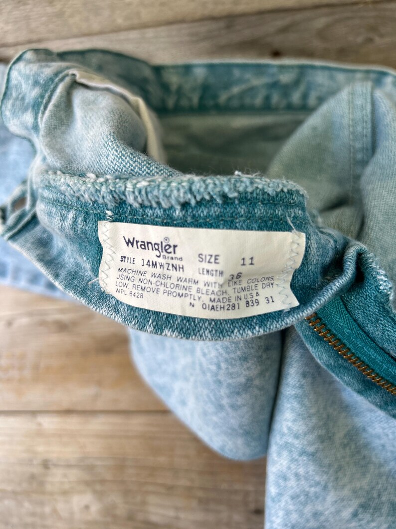 May include: Light blue acid-wash denim jeans featuring a white interior tag with the Wrangler brand logo, style number 14MWZNH, size 11, and length 36. The tag includes care instructions for machine washing and tumble drying, and states Made in USA.