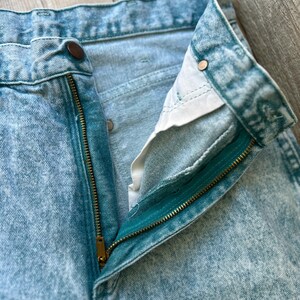 May include: High-waisted denim jeans in a light blue acid wash finish. The jeans feature a metal zipper fly, a single button closure at the waistband, and copper-toned metal rivets at the pocket corners. The interior pocket lining is white.