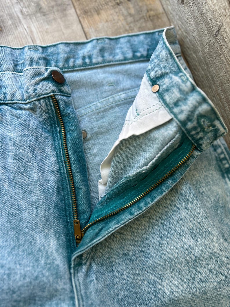 May include: High-waisted denim jeans in a light blue acid wash finish. The jeans feature a metal zipper fly, a single button closure at the waistband, and copper-toned metal rivets at the pocket corners. The interior pocket lining is white.