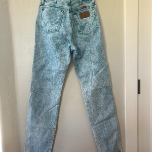 May include: High-waisted acid wash denim jeans in a light blue and white mottled pattern. The jeans feature two back pockets with a signature W-stitch design, copper-toned rivets, a brown leather patch on the waistband, and a button fly closure.