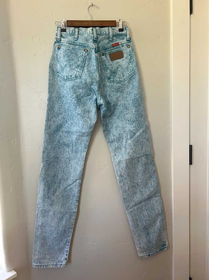 May include: High-waisted acid wash denim jeans in a light blue and white mottled pattern. The jeans feature two back pockets with a signature W-stitch design, copper-toned rivets, a brown leather patch on the waistband, and a button fly closure.