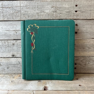 May include: A dark green photo album with a gold floral design in the upper left corner and a gold border. The album is closed and appears to be made of a textured fabric. The background is a wooden surface.