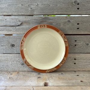 May include: A beige ceramic plate with a brown rim and decorative circular patterns. The plate is round and appears to be vintage or retro in style. The plate is sitting on a wooden surface.