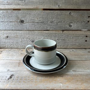 May include: A white ceramic coffee cup with a brown rim and handle sits on a matching saucer. The saucer has a black and white striped border. The set is on a wooden surface.