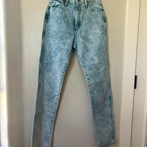 May include: High-waisted jeans in a light blue acid wash denim finish. These pants feature a classic five-pocket design, a button and zipper fly closure, and copper-toned rivets at the pocket corners. The denim has a mottled, textured appearance.