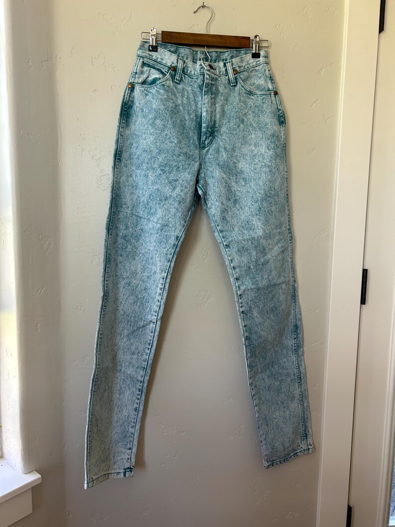 May include: High-waisted jeans in a light blue acid wash denim finish. These pants feature a classic five-pocket design, a button and zipper fly closure, and copper-toned rivets at the pocket corners. The denim has a mottled, textured appearance.