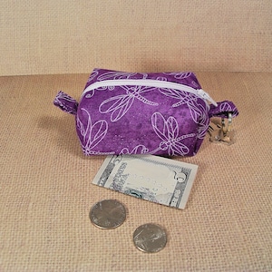 May include: A small purple zippered pouch with a white dragonfly pattern. The pouch is sitting on a brown burlap surface with a five dollar bill and two coins.