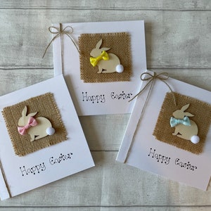 May include: Three white square Easter cards with a burlap square, wooden bunny, and a bow. Each card has a different coloured bow: pink, yellow, and blue. The words "Happy Easter" are printed below the burlap square.
