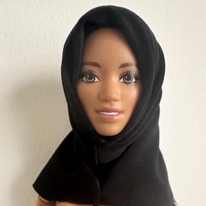 May include: A doll with brown skin and long eyelashes wearing a black hijab. The doll's face is centered in the frame, with the black fabric of the hijab framing the face and shoulders. The background is a plain white wall.