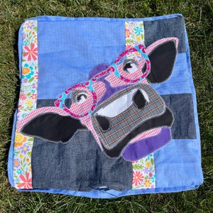 May include: A blue and denim patchwork pillow with a colorful applique of a cow's face wearing glasses. The cow's face is made of different fabrics and has a purple nose and black eyes.