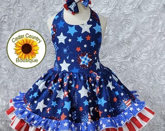 Op de afbeelding: Een blauwe en rode, mouwloze jurk met een witte sterren- en vuurwerkprint. De jurk heeft een geruite rok met rode, witte en blauwe strepen. De jurk wordt gecombineerd met een rood, wit en blauw strikband. De jurk is gelabeld met "Cedar Country Boutique".