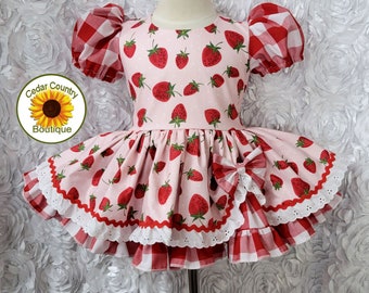 May include: A pink and red strawberry print dress with a white lace trim and a red and white gingham bow. The dress has a puffed sleeve and a full skirt.