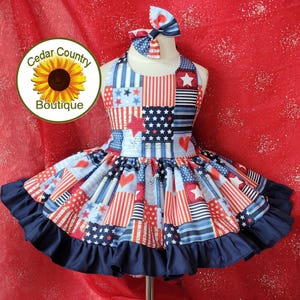 May include: A sleeveless dress featuring a patchwork pattern of red, white, and blue stripes, stars, and hearts, finished with a solid navy blue ruffled hem. The dress includes a matching hair bow with a similar patriotic star and stripe design.