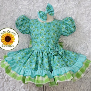 May include: A turquoise dress with a frog pattern, ruffled skirt, and matching bow. The dress features a turquoise and green plaid trim and a bow at the waist. The dress is designed for a child.