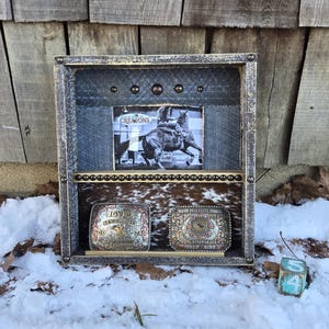 May include: A rustic wooden shadow box with a black and white image of a horse and rider. The box features two belt buckles and a cowhide shelf. The word "CREATIONS" is visible on the image.