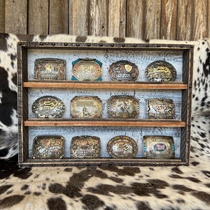 May include: A wooden display case with three shelves, each holding a row of belt buckles. The buckles are silver and gold with various designs and inscriptions. The case has a rustic, distressed look with metal accents.