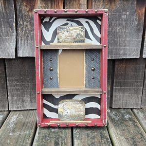 May include: A rustic, red-framed display shelf with a zebra-print backing. It features two shelves and a central space for a photo or item. Two belt buckles are displayed, one on each shelf, with text and decorative details.