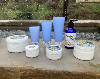 May include: A set of six white jars and tubes of hand cream with blue labels that say "Pudlani Beauty". The jars and tubes are arranged on a gray stone surface. The background is a blurry image of a pond and green foliage.