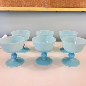May include: Six light blue glass dessert bowls. Each bowl has a rounded cup shape, a ball-shaped stem, and a circular base. The bowls are arranged on a white surface, with a light blue background.