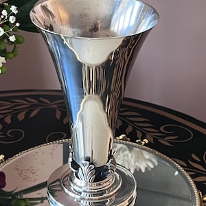 May include: A silver-colored vase with a flared top and a decorative base. The vase has a smooth, reflective surface and a detailed, ornate design at the base. The vase is likely made of metal and is suitable for displaying flowers or as a decorative item.