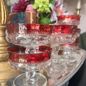 May include: A collection of vintage dessert glasses with ruby red rims and clear, patterned bowls. The glasses are displayed on a silver serving tray, showcasing their elegant design. The background features a floral arrangement.