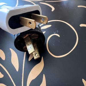 K&ouml;nnte beinhalten: Nahaufnahme von zwei elektrischen Steckern. Einer ist wei&szlig; mit der Aufschrift "15A 125V", der andere ist schwarz. Beide haben goldfarbene Stifte. Der Hintergrund ist eine schwarze Oberfl&auml;che mit einem goldenen Blumenmuster.