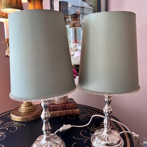 K&ouml;nnte beinhalten: Zwei passende Tischlampen mit silberfarbenen Sockeln und hellgr&uuml;nen Schirmen. Die Schirme sind zylindrisch und mit einem Perlenrand versehen. Die Lampen stehen auf einem dunkelblauen Tisch mit goldfarbenen floralen Mustern.