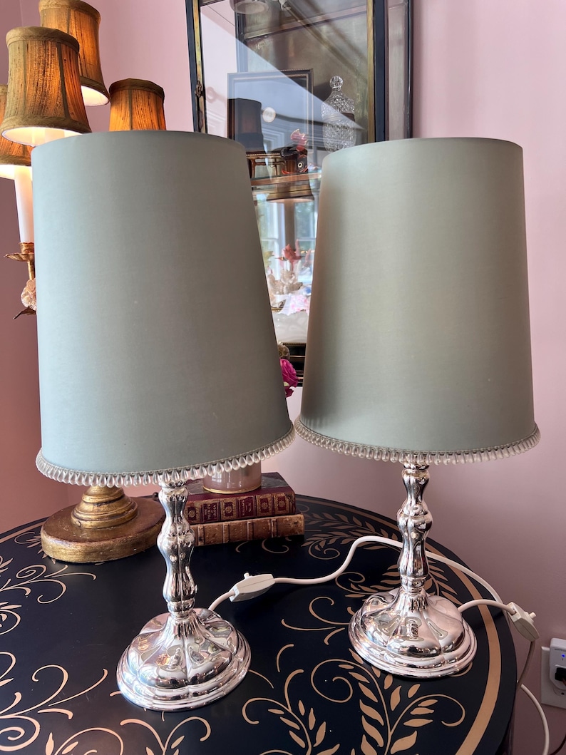 K&ouml;nnte beinhalten: Zwei passende Tischlampen mit silberfarbenen Sockeln und hellgr&uuml;nen Schirmen. Die Schirme sind zylindrisch und mit einem Perlenrand versehen. Die Lampen stehen auf einem dunkelblauen Tisch mit goldfarbenen floralen Mustern.