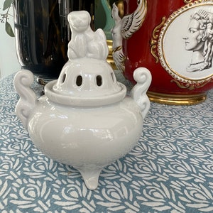 May include: White ceramic incense burner with a lid shaped like a sitting cat. The burner has four legs and a round body with holes for incense sticks. The burner is sitting on a blue and white patterned surface.