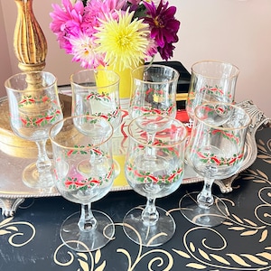 May include: A set of six clear glass goblets with gold rims, each decorated with a painted pattern of green holly leaves and red ribbons. The glasses feature a short stem and a round base, arranged in a group on a reflective silver tray.