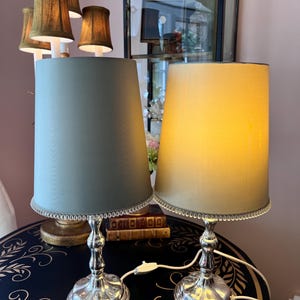 K&ouml;nnte beinhalten: Zwei elegante Tischlampen mit silbernen Sockeln und konischen Schirmen. Ein Schirm ist in ged&auml;mpftem Gr&uuml;n, der andere in weichem Gelb gehalten. Beide Schirme sind mit einem silbernen Perlenrand versehen. Die Lampen stehen auf einem dunklen Tisch mit floralem Design.