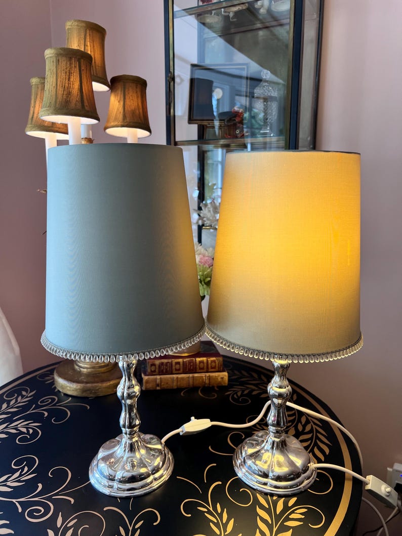 K&ouml;nnte beinhalten: Zwei elegante Tischlampen mit silbernen Sockeln und konischen Schirmen. Ein Schirm ist in ged&auml;mpftem Gr&uuml;n, der andere in weichem Gelb gehalten. Beide Schirme sind mit einem silbernen Perlenrand versehen. Die Lampen stehen auf einem dunklen Tisch mit floralem Design.