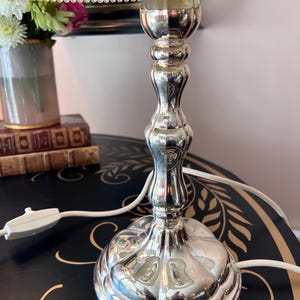 K&ouml;nnte beinhalten: Eine silberfarbene Tischlampe mit dekorativem Fu&szlig; und Schaft. Die Lampe hat ein wei&szlig;es Kabel und einen hellen Lampenschirm mit Perlenbesatz. Eine Vase mit Blumen und B&uuml;cher befinden sich im Hintergrund.