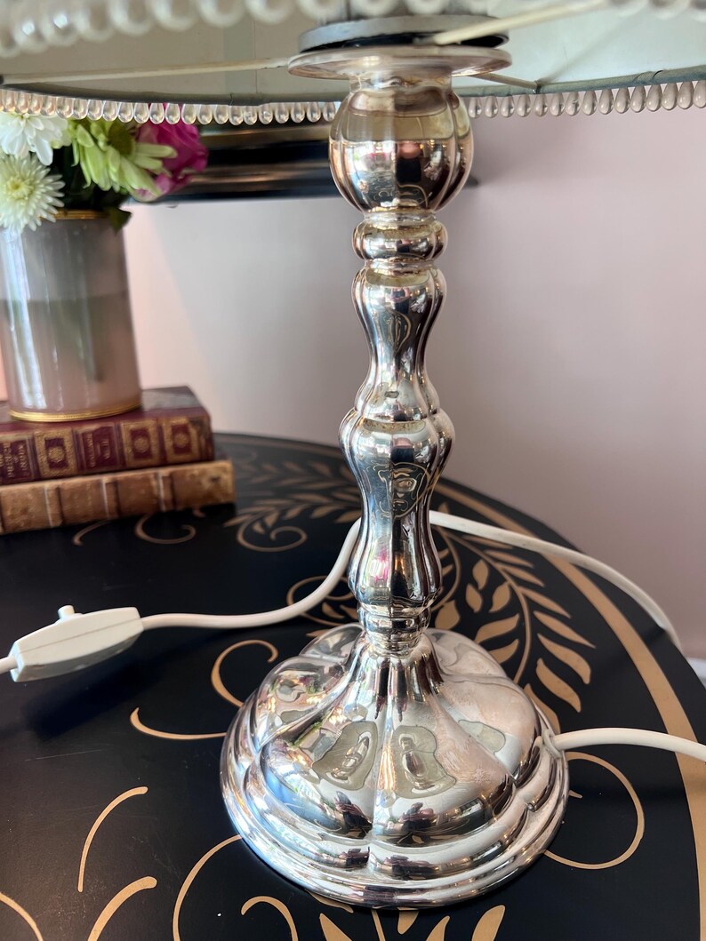 K&ouml;nnte beinhalten: Eine silberfarbene Tischlampe mit dekorativem Fu&szlig; und Schaft. Die Lampe hat ein wei&szlig;es Kabel und einen hellen Lampenschirm mit Perlenbesatz. Eine Vase mit Blumen und B&uuml;cher befinden sich im Hintergrund.