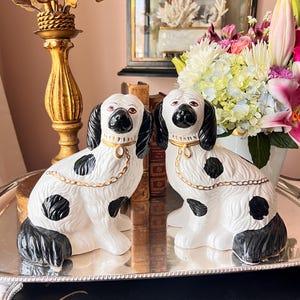 May include: Pair of ceramic dog figurines. The dogs are white with black spots, ears, and noses. They have gold-colored collars and chains. The dogs are sitting on a silver tray, with a blurred background of flowers and a mirror.