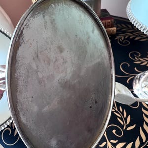 K&ouml;nnte beinhalten: Ein ovales Tablett aus silberfarbenem Metall mit leicht angelaufener Oberfl&auml;che. Das Tablett hat einen erh&ouml;hten Rand und ein schlichtes, elegantes Design. Der Hintergrund umfasst eine dunkelblaue Oberfl&auml;che mit einem goldenen Bl&auml;ttermuster und eine Blumenvase.