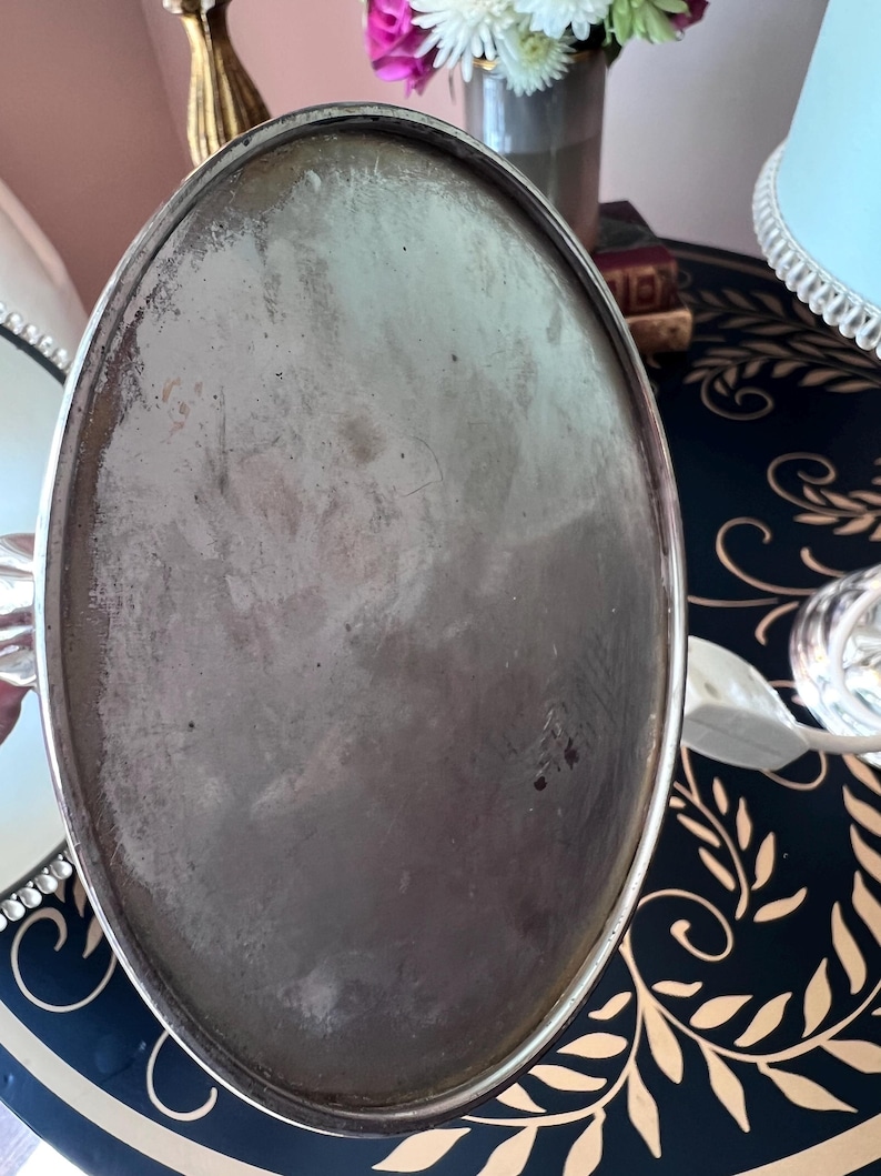 K&ouml;nnte beinhalten: Ein ovales Tablett aus silberfarbenem Metall mit leicht angelaufener Oberfl&auml;che. Das Tablett hat einen erh&ouml;hten Rand und ein schlichtes, elegantes Design. Der Hintergrund umfasst eine dunkelblaue Oberfl&auml;che mit einem goldenen Bl&auml;ttermuster und eine Blumenvase.