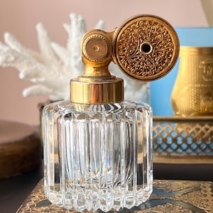 May include: A vintage perfume bottle with a clear, ribbed glass body and a gold-toned atomizer. The atomizer features an ornate, circular design. The bottle is displayed on a decorative surface, suggesting a focus on antique or decorative items.