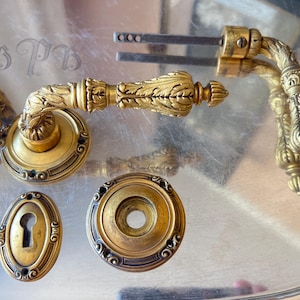 May include: Antique brass door hardware set, including a handle, escutcheon, and keyhole cover. The pieces feature ornate floral and leaf detailing, with a warm, aged patina. The set is displayed on a silver tray.