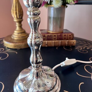 K&ouml;nnte beinhalten: Eine silberne Tischlampe mit geriffeltem Fu&szlig; und hellblauem Lampenschirm. Die Lampe steht auf einer dunklen Oberfl&auml;che mit floralem Design. Eine goldene Lampe und eine Blumenvase befinden sich im Hintergrund.