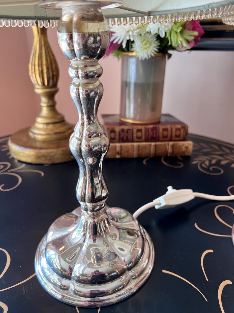 K&ouml;nnte beinhalten: Eine silberne Tischlampe mit geriffeltem Fu&szlig; und hellblauem Lampenschirm. Die Lampe steht auf einer dunklen Oberfl&auml;che mit floralem Design. Eine goldene Lampe und eine Blumenvase befinden sich im Hintergrund.