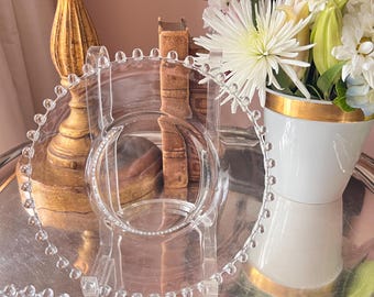 May include: A clear glass serving dish with a beaded edge. The dish is on a silver tray, with a white vase of flowers and a gold candlestick in the background. The flowers are pink and white.
