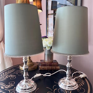 K&ouml;nnte beinhalten: Zwei silberne Tischlampen mit salbeigr&uuml;nen Schirmen. Die Lampen haben verzierte silberne Sockel und einen Perlenbesatz. Sie stehen auf einem dunkelblauen Tisch mit goldenem Blattmuster.