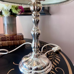 K&ouml;nnte beinhalten: Silberne Tischlampe mit dekorativem Fu&szlig; und hellblauem Lampenschirm. Die Lampe hat ein verziertes Design und ein wei&szlig;es Netzkabel. Eine Vase mit Blumen befindet sich im Hintergrund.