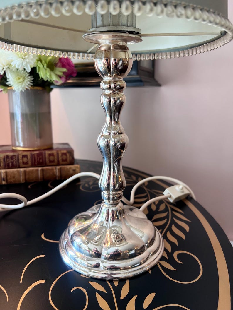 K&ouml;nnte beinhalten: Silberne Tischlampe mit dekorativem Fu&szlig; und hellblauem Lampenschirm. Die Lampe hat ein verziertes Design und ein wei&szlig;es Netzkabel. Eine Vase mit Blumen befindet sich im Hintergrund.