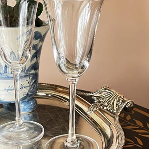 May include: Two clear, stemmed wine glasses with a fluted design, resting on a silver tray. The glasses have a delicate, elegant appearance, with a slender stem and a wide, tulip-shaped bowl. A blue and white vase is in the background.