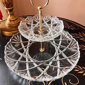 May include: A two-tiered, clear glass serving tray with a gold-colored handle. The glass has a diamond-cut pattern. The top tier is smaller than the bottom tier. The handle is a gold-colored metal. The serving tray is likely used for displaying desserts or appetizers.