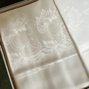 May include: Ivory-colored tablecloth with a detailed floral pattern, neatly folded inside a box. The design features elegant, swirling leaves and vines, creating a classic and sophisticated look. The fabric has a subtle sheen.