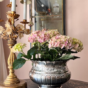 May include: A silver-colored, ornate planter filled with pink and green hydrangeas. The planter sits on a black table with gold accents, next to a gold candelabra. A mirrored cabinet is in the background.