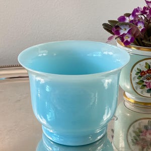May include: A light blue glass planter with a rounded base and flared rim. The planter is sitting on a silver tray. A second, decorated planter with flowers is visible in the background.